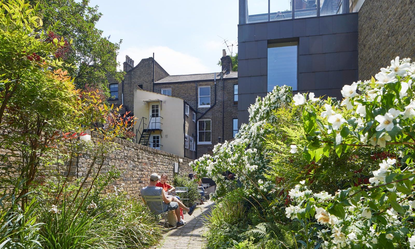 This photo is a view of the SLG's Fox Garden, it shows bushes and plants in the foreground, a person sitting on a chair in the mid-ground and the building of the gallery in the background.