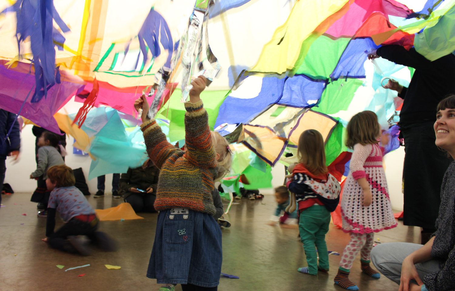 A small child raises her hands to the air to touch lots of pieces of coloured tissue paper above her. Behind other children are playing. To the right of the picture going out of the shot a woman is crouching down and smiling.