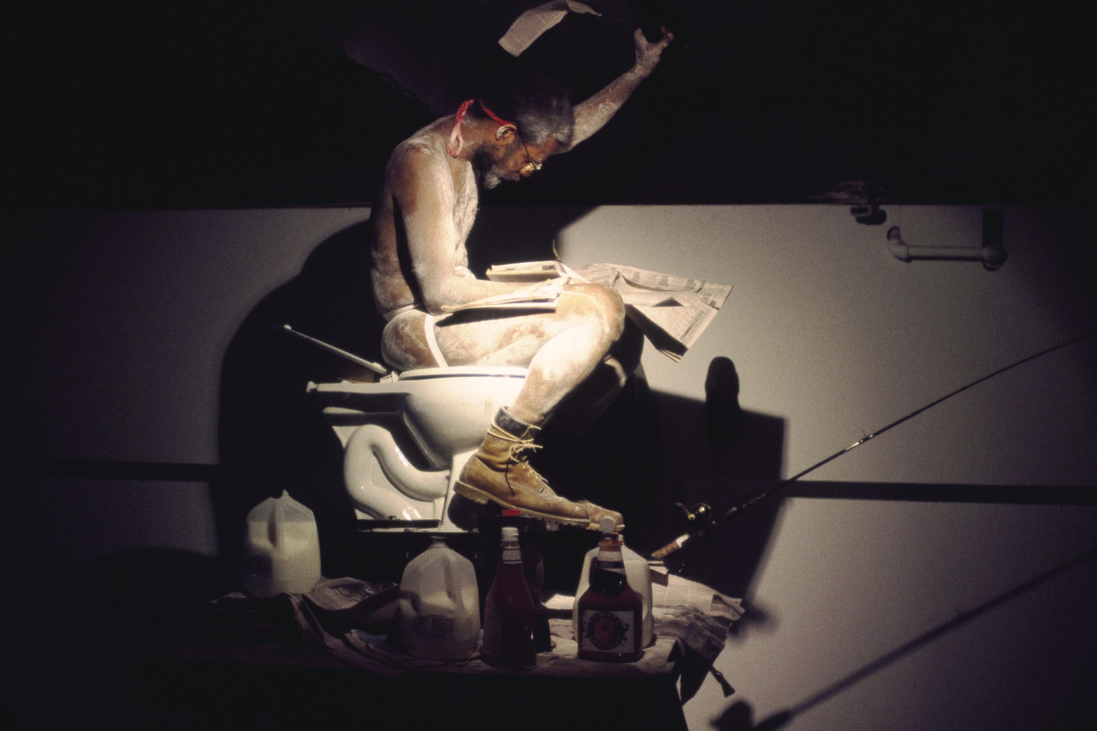 A man sitting on a toilet wearing a jockstrap and covered in flour, tearing pages from a newspaper