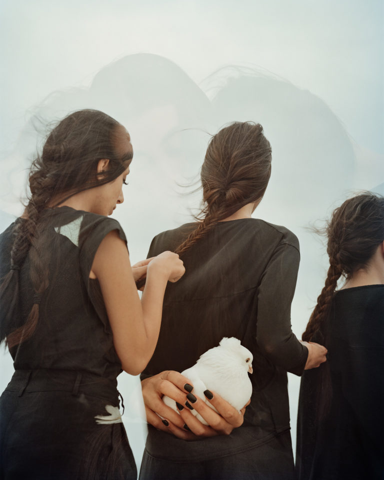 Three woman stand in a line and braid each others hair. They are wearing all black. Overlaid on this image is a photo of hands holding a white dove. This is an artwork by Hoda Afshar.