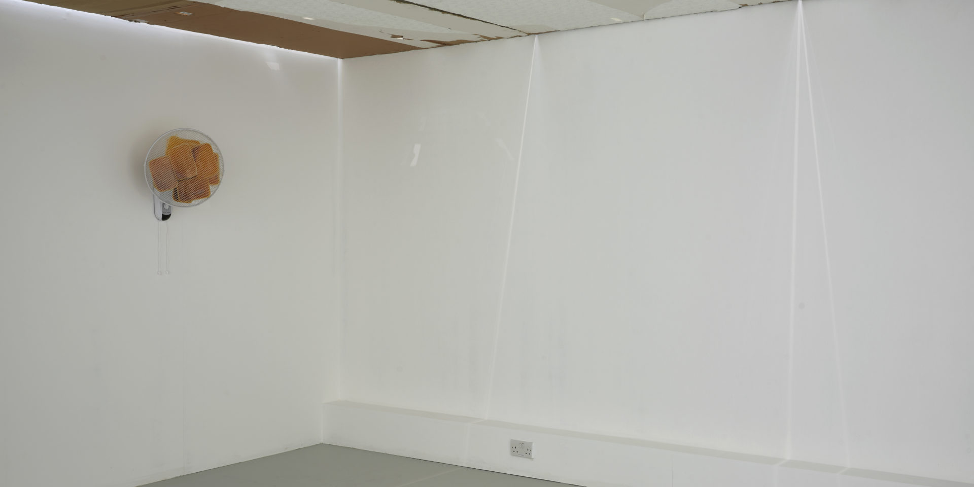 A white gallery room with art installed. The art is mixed media and includes a fan installed on the wall.