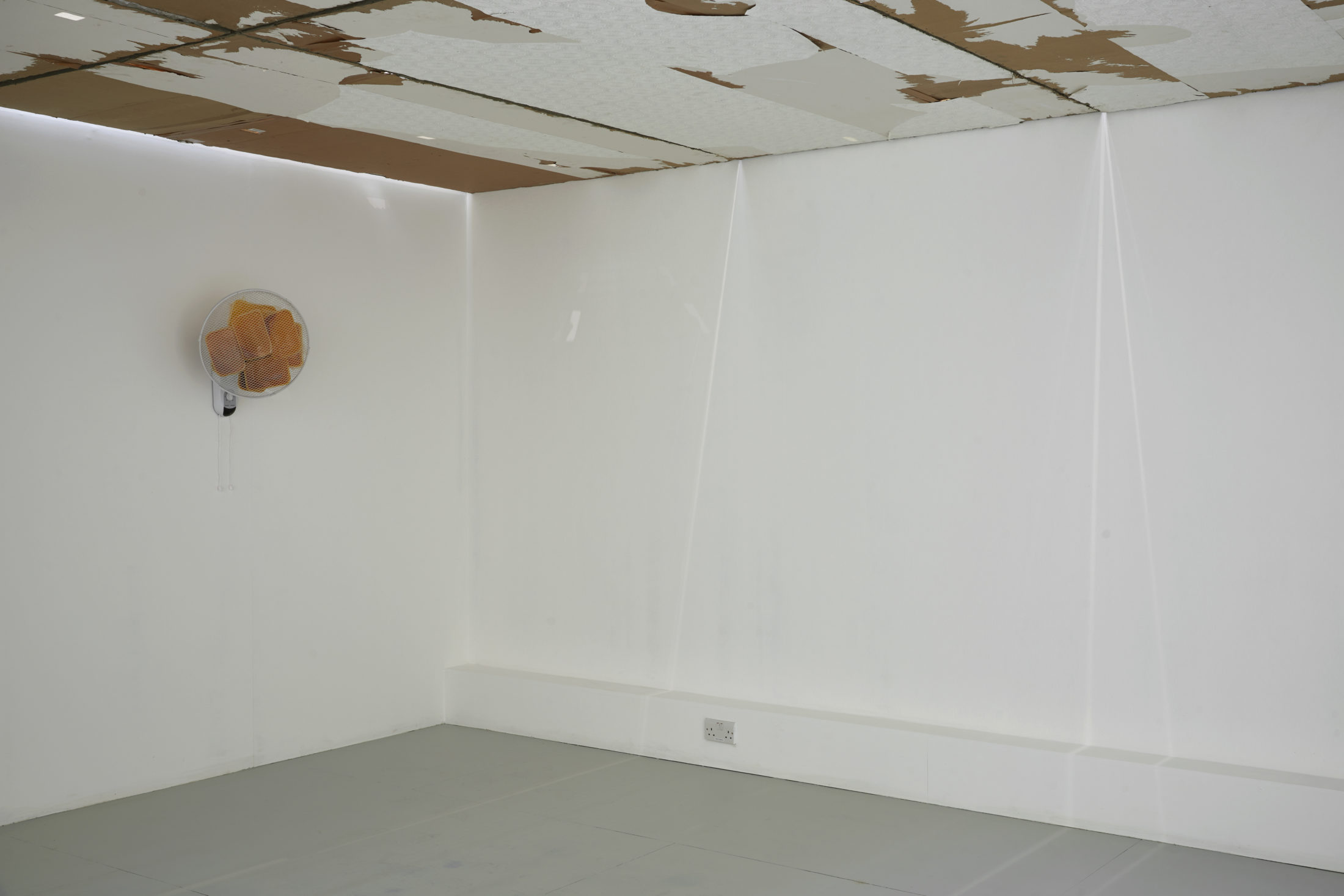 A white gallery room with art installed. The art is mixed media and includes a fan installed on the wall.