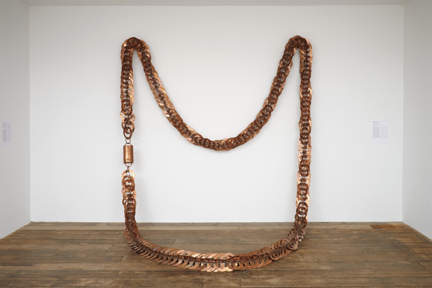 A large copper necklace sculpture hanging on a gallery wall. The work is by artist Ana Pellicer.